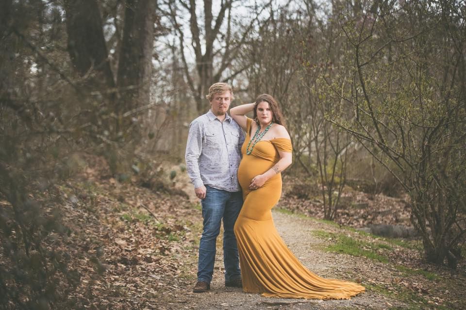 Couple maternity photo on forest path, woman in golden yellow gown with partner in casual attire.