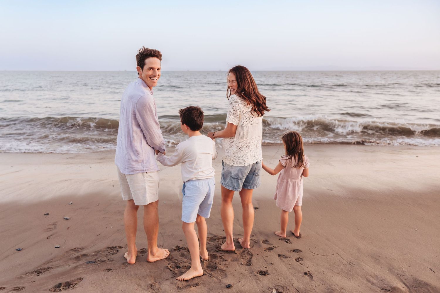 Family of four holding hands on a sandy beach at sunset, with ocean waves in the background, what to wear to beach photo session