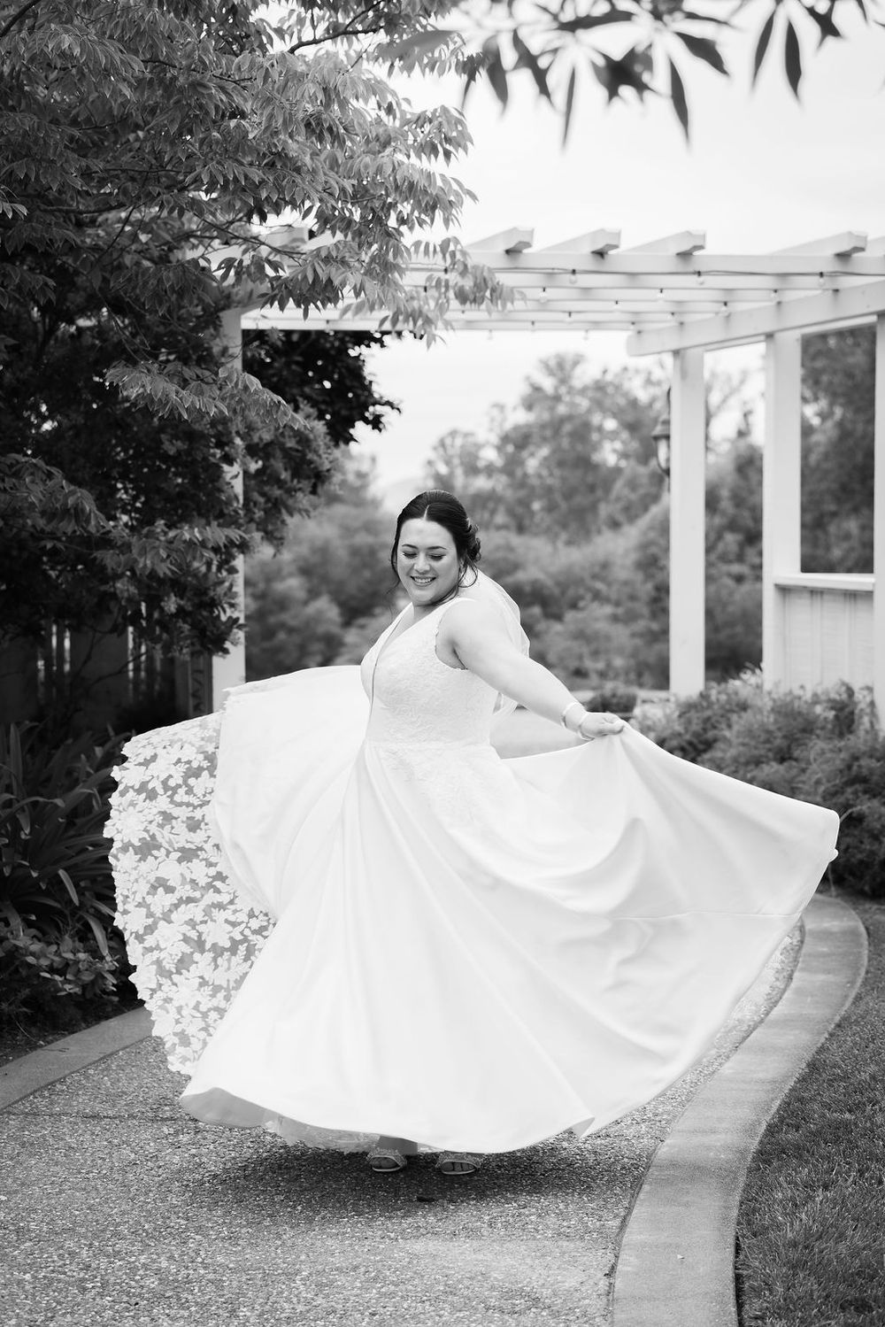 Bride in white lace gown twirling on garden path in black and white wedding photo.