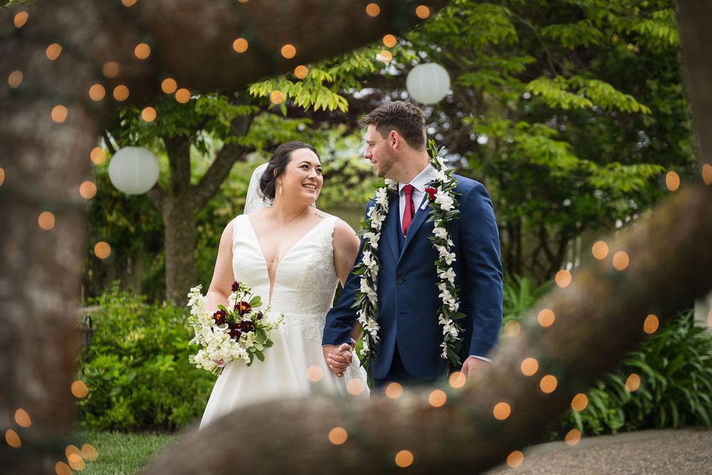 Bride and groom holding hands in a garden, surrounded by string lights and greenery, groom wearing a floral lei.