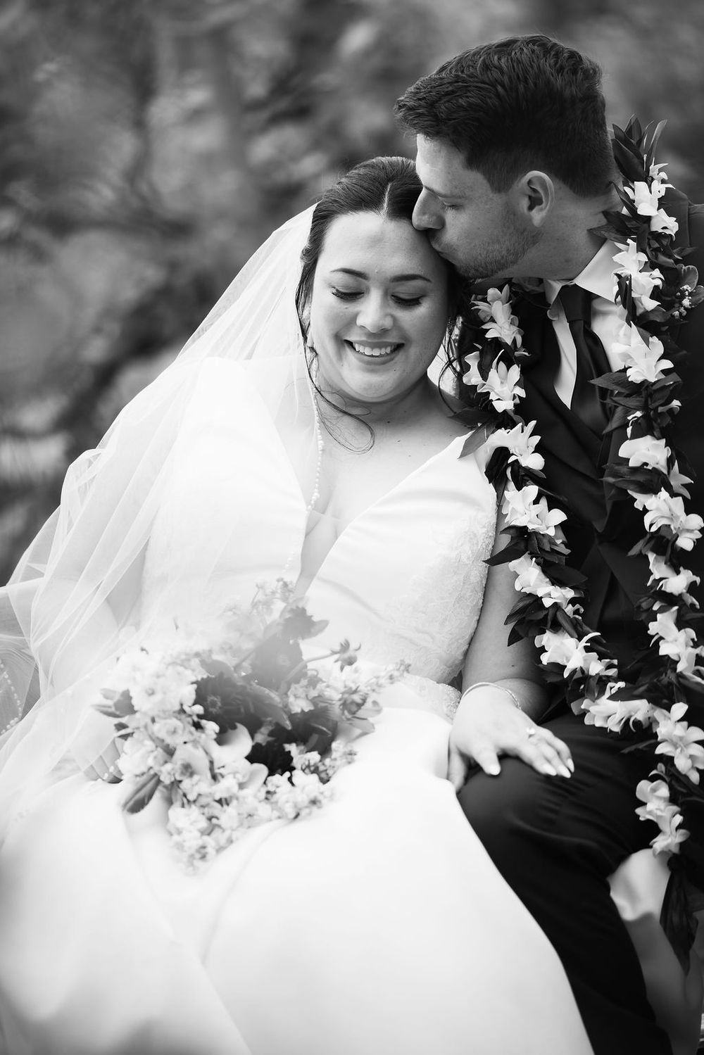 Black and white photo of a smiling bride holding bouquet while groom kisses her cheek, both wearing floral leis.