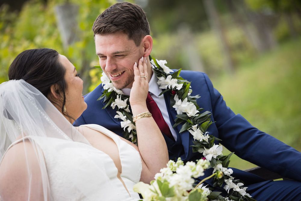 Bride and groom share a romantic moment sitting outdoors, groom wearing a floral lei and navy suit.