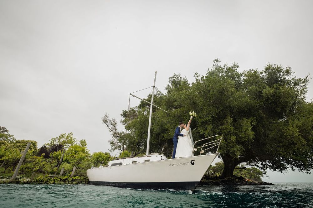 Bride and groom kissing on a white sailboat surrounded by lush green trees on calm water.