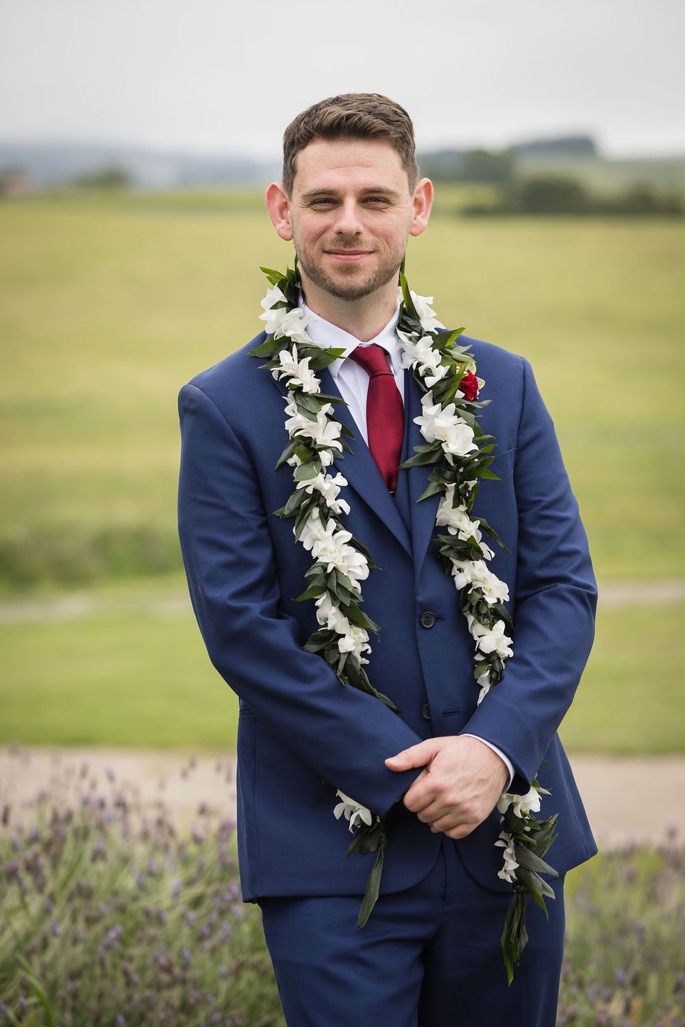 Groom in navy blue suit wearing white floral lei with red tie at outdoor wedding.