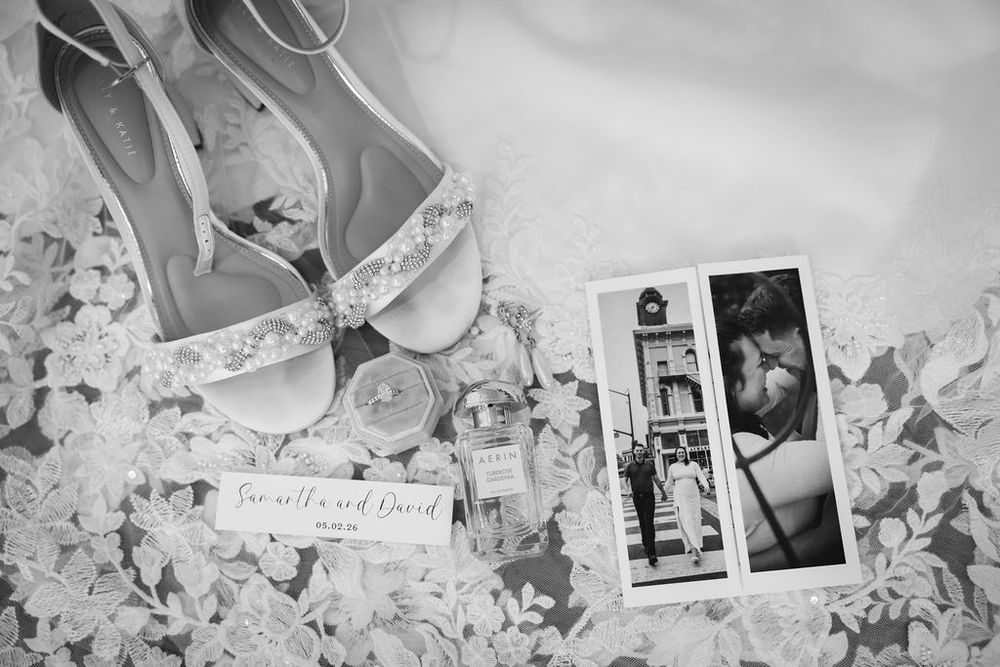 Black and white flat lay of bridal accessories including shoes, perfume, and wedding invitation.