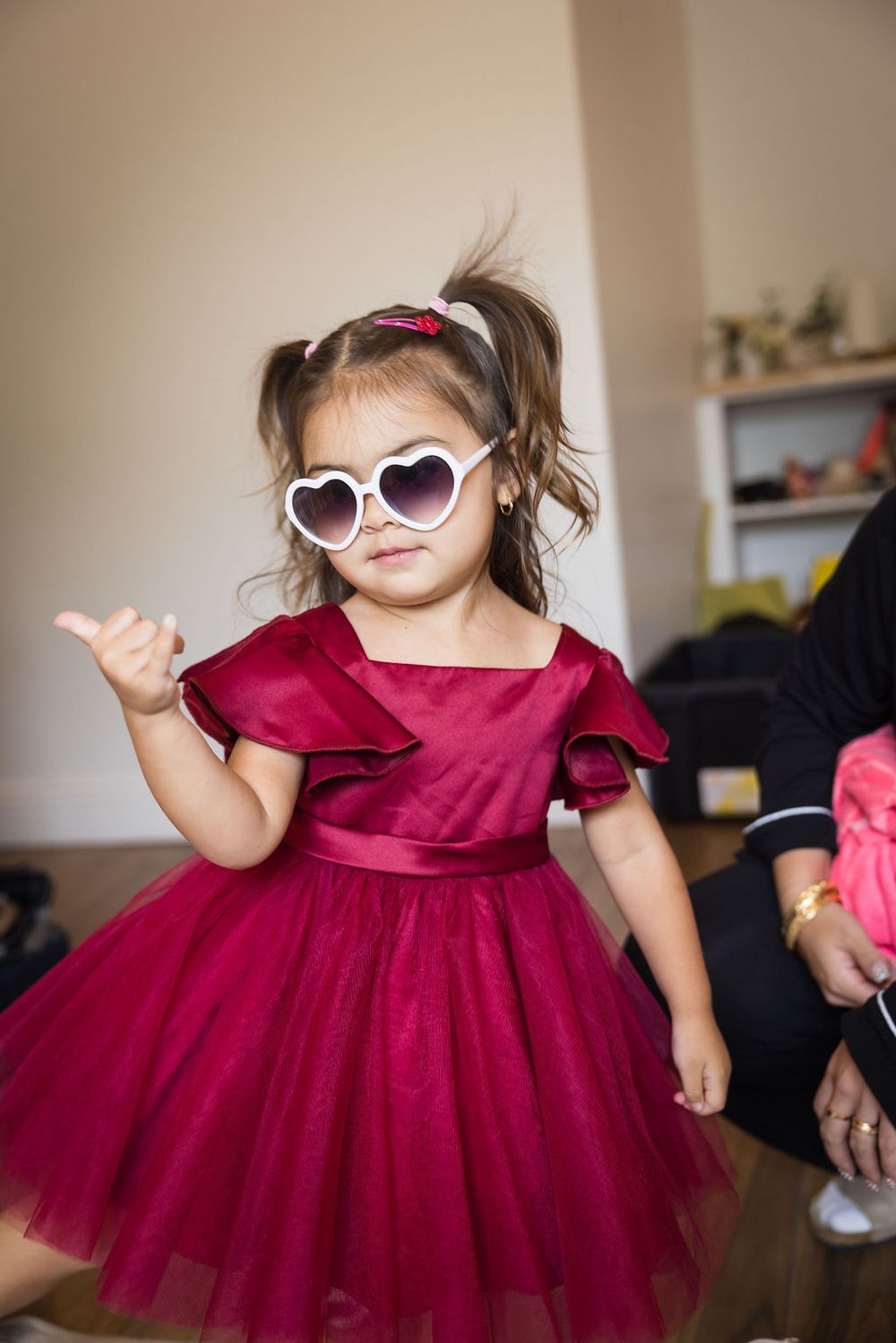 Adorable toddler girl in a crimson tulle dress and heart-shaped sunglasses posing confidently indoors.