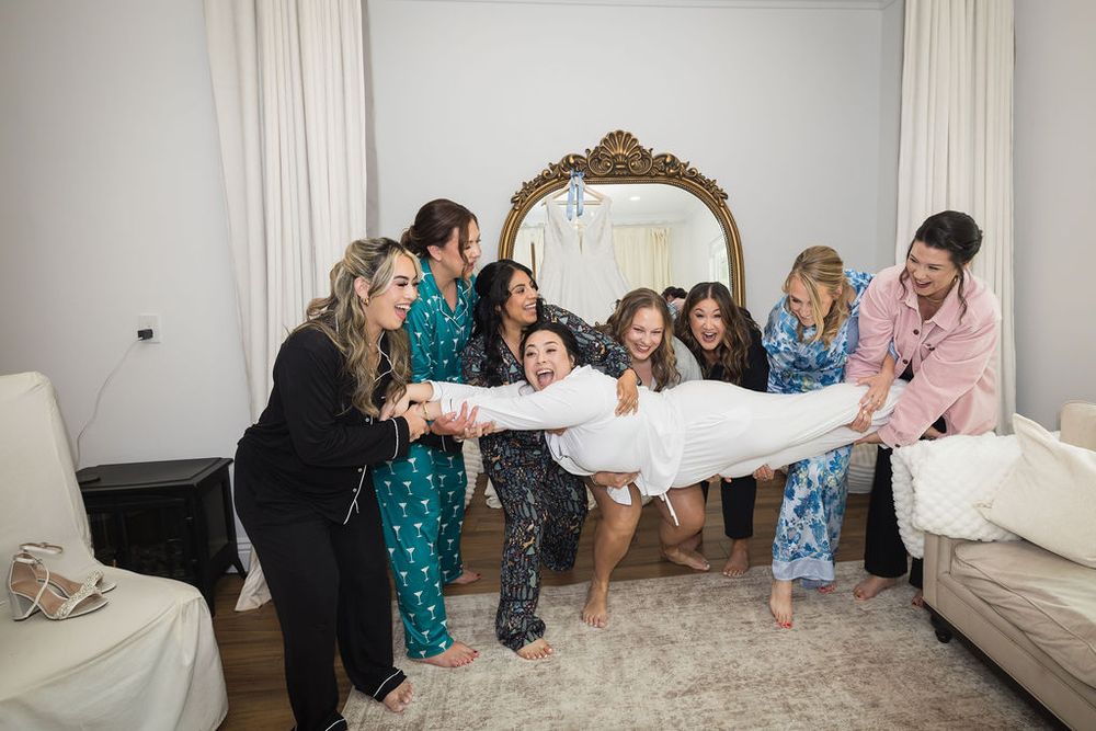 Bridesmaids in pajamas playfully lifting bride during fun wedding morning getting ready party.
