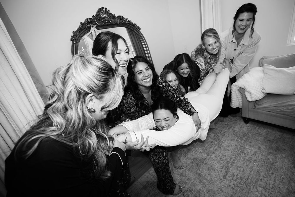 Bridesmaids playfully lift the bride during wedding preparations in a black and white photo.