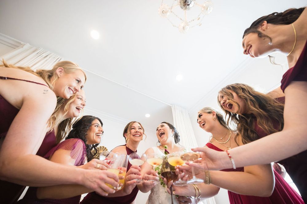 Bridesmaids in burgundy dresses toasting with champagne glasses in joyful low-angle wedding photo.