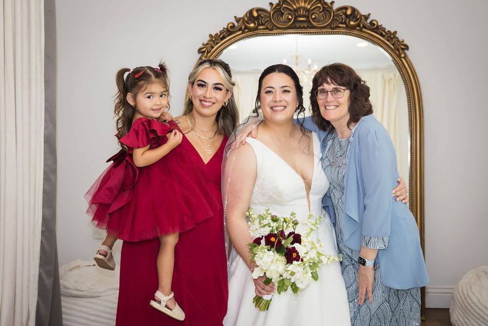 Bride in white gown holds bouquet with bridesmaid in red dress holding child and older woman in blue at wedding.