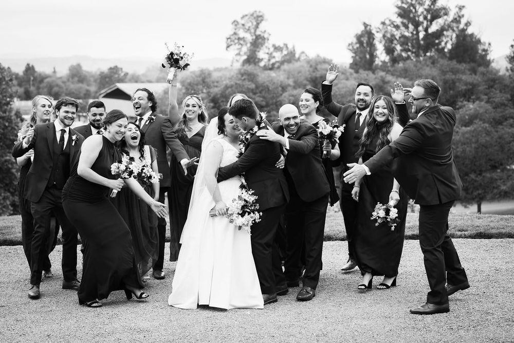 Black and white photo of a joyful wedding party celebrating as the couple shares a kiss.