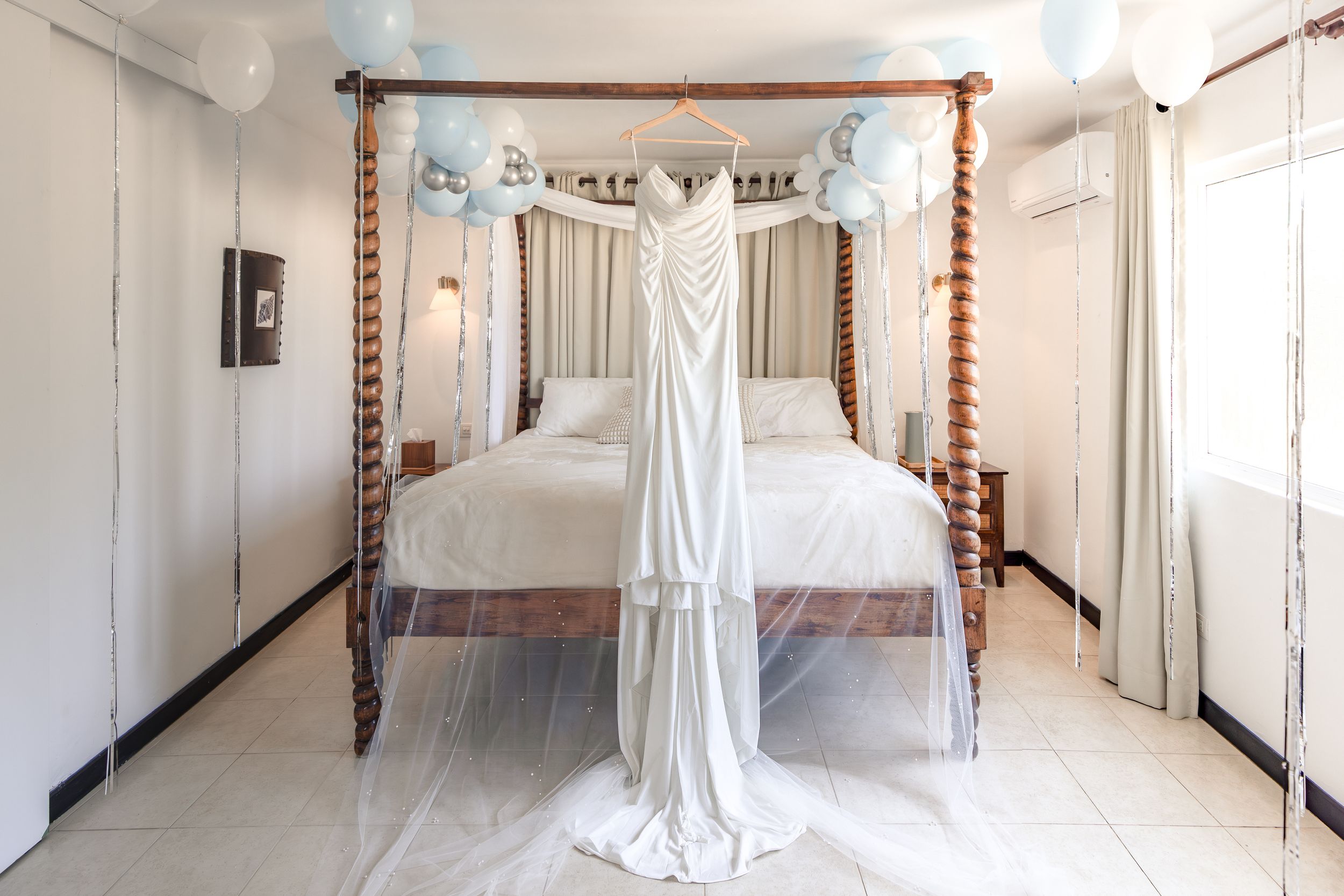 White wedding dress hanging on a four-poster bed decorated with blue balloons in an elegant white bedroom.