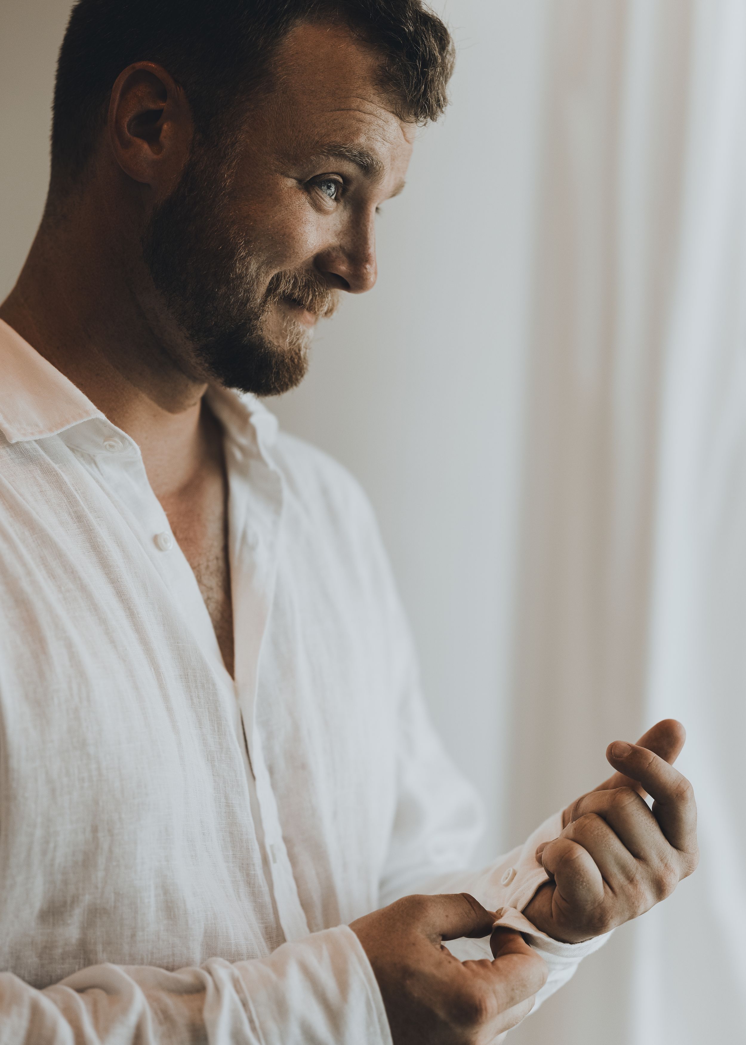 Smiling bearded man in white linen shirt adjusting cufflinks near window.