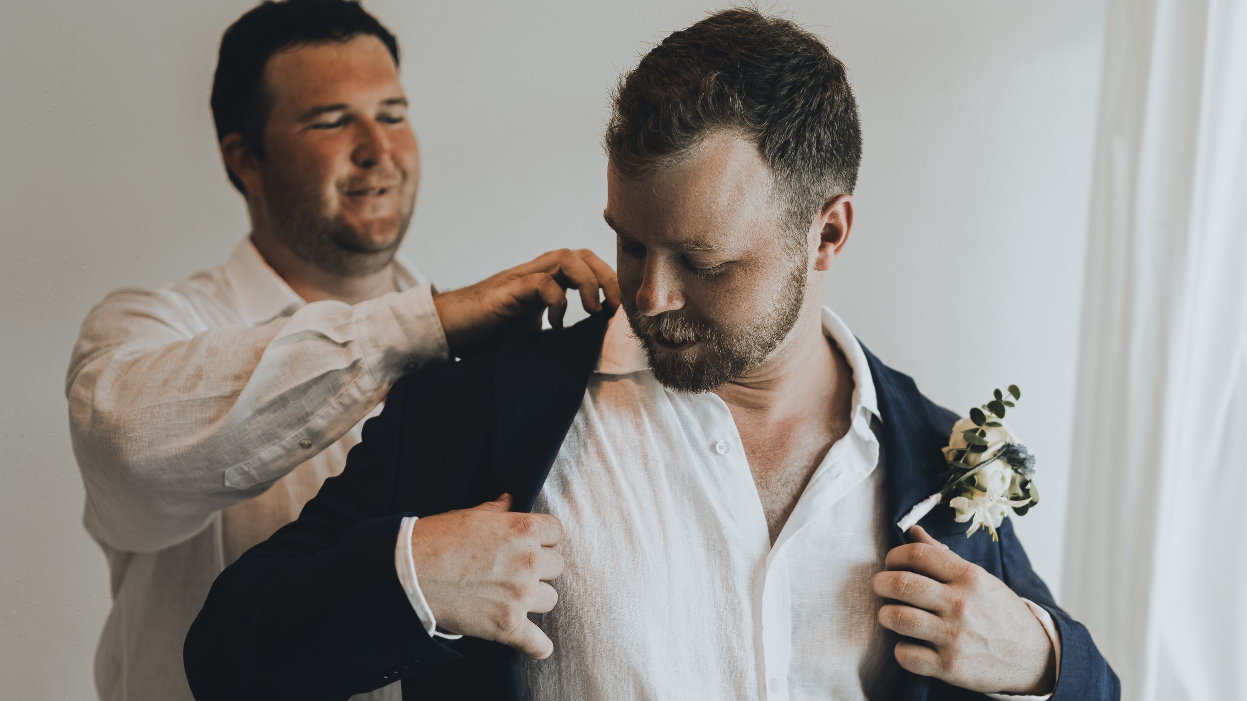 A groomsman helps a groom in a navy suit adjust his jacket before a wedding ceremony.