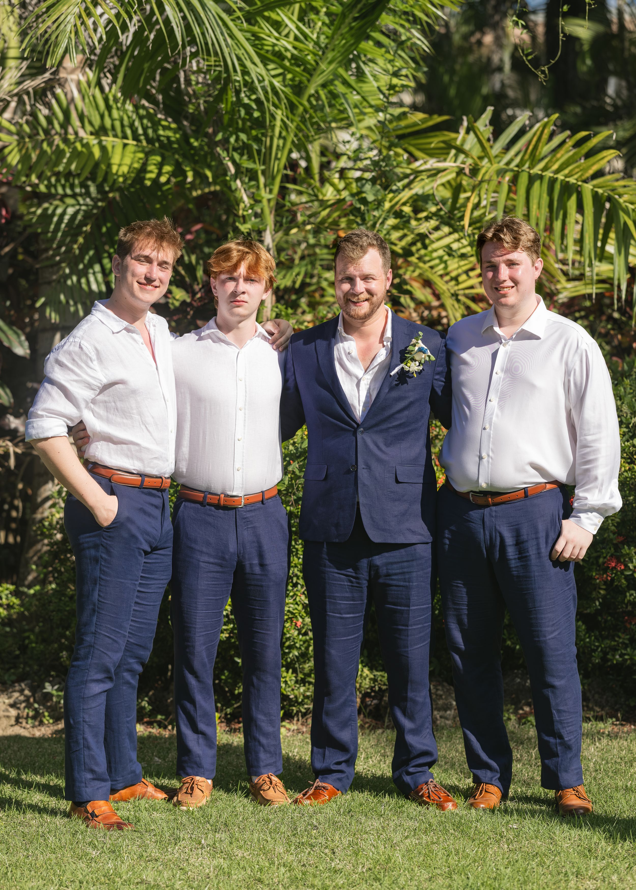 Groom in navy suit stands with three groomsmen in white shirts and navy pants outdoors at wedding.