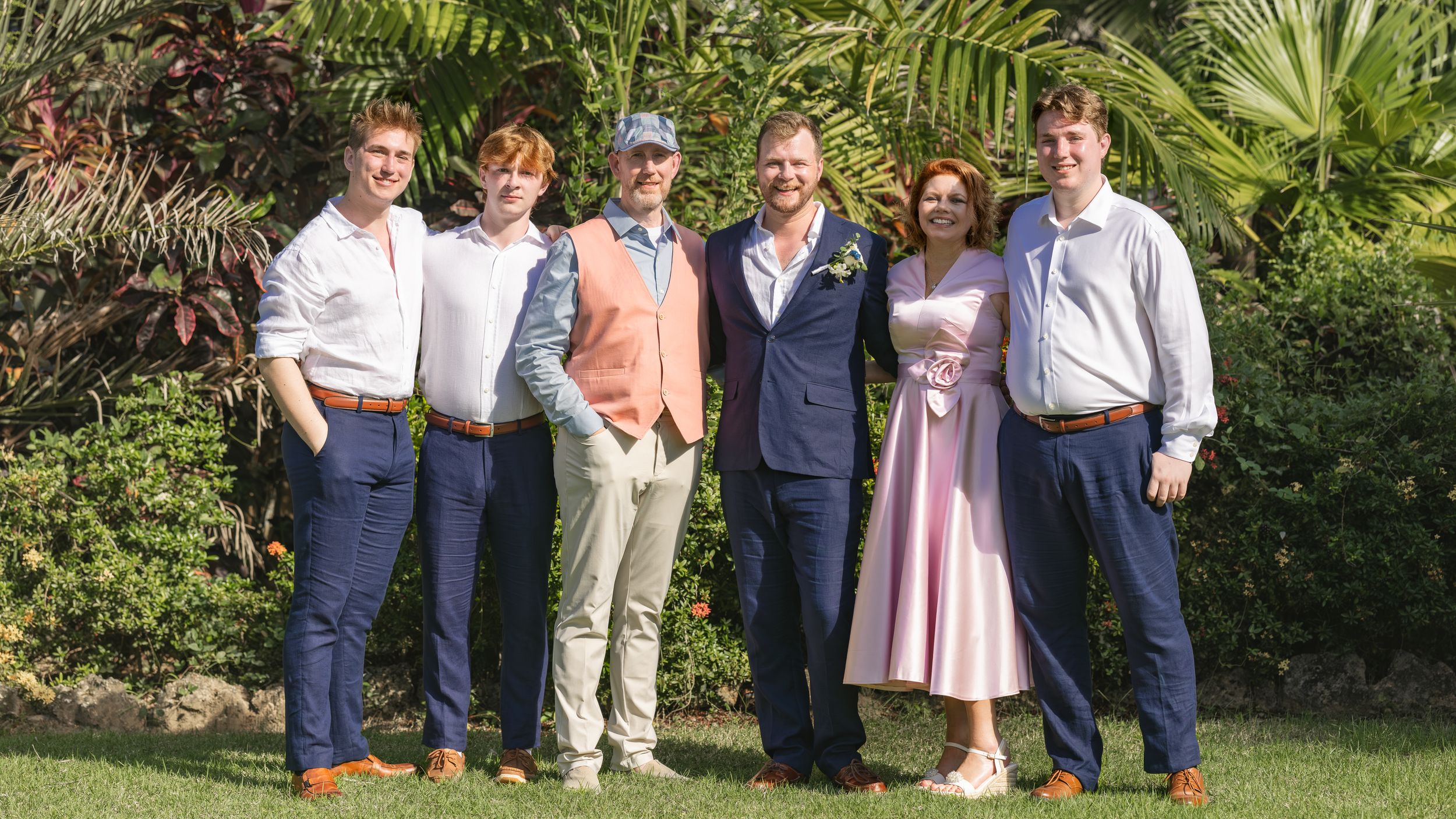 Group of six people posing outdoors at a tropical wedding celebration, wearing formal attire including pink dress and navy suits.