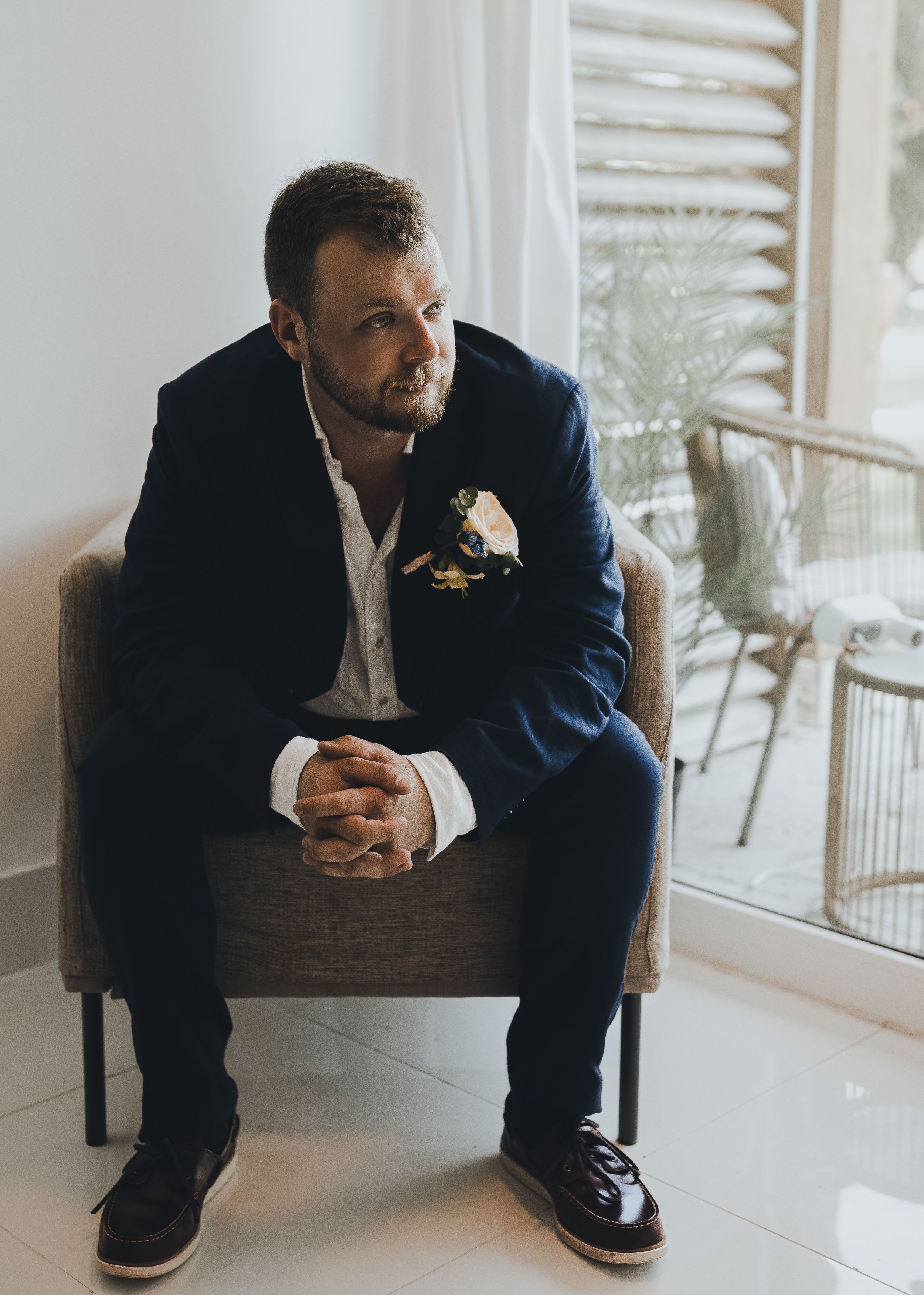 Stylish bearded man in navy suit with boutonniere sitting in chair by window.