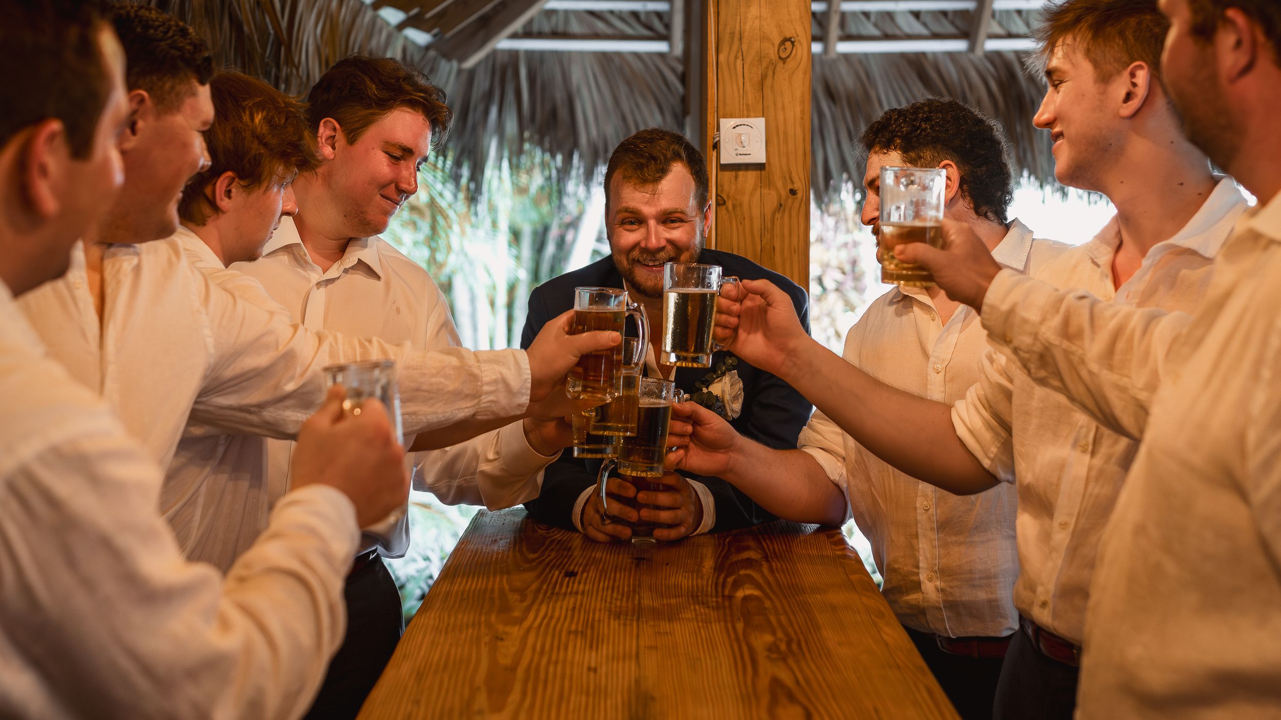Group of young men in white shirts toasting with beer mugs at a wooden bar table in a festive celebration.