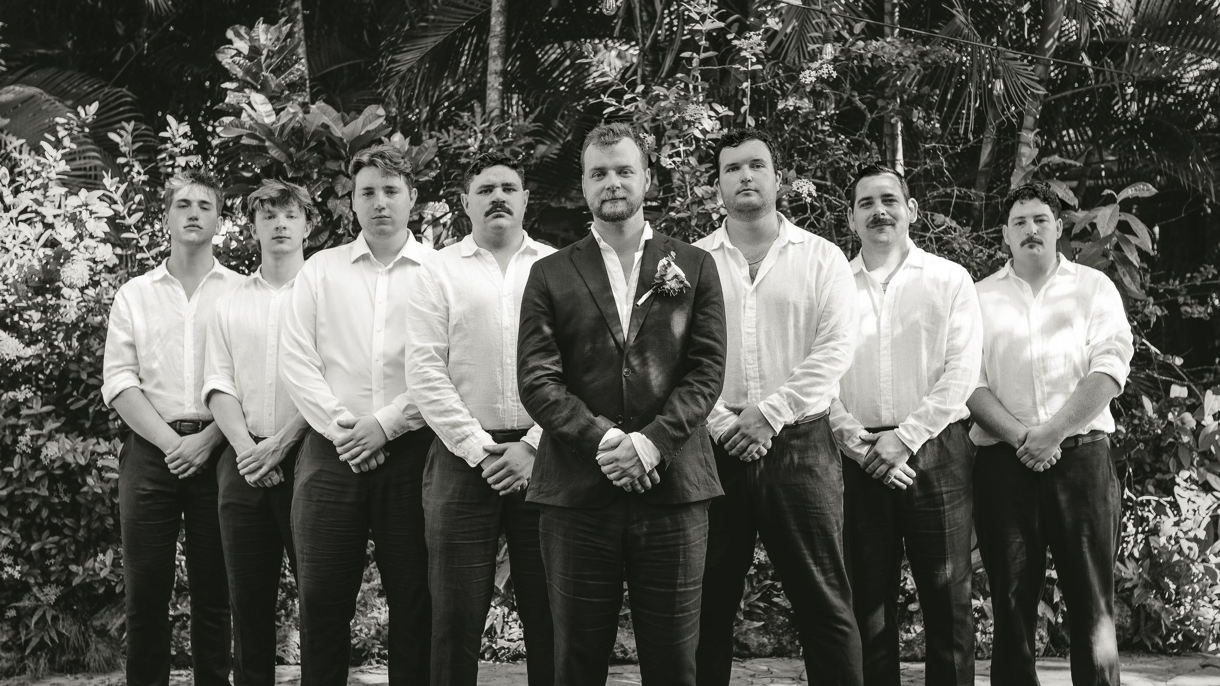 Black and white photo of a groom in a suit with groomsmen in white shirts outdoors.