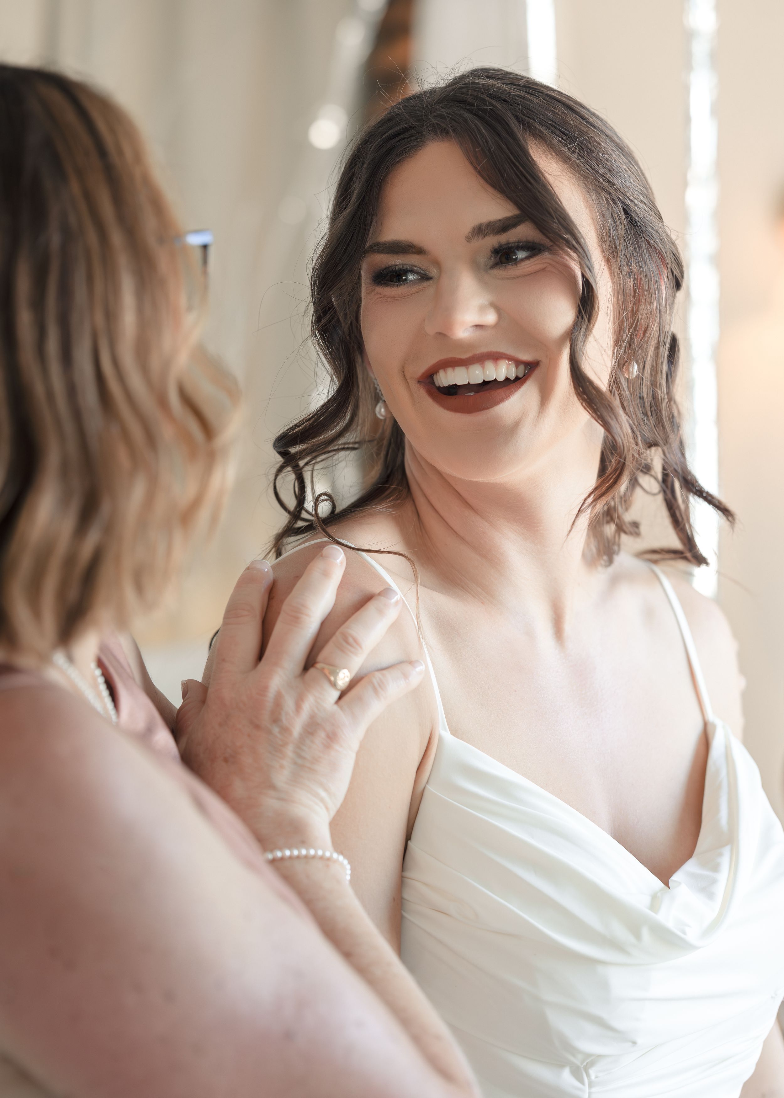 Smiling bride in white gown laughing with older woman touching her shoulder during wedding preparations.