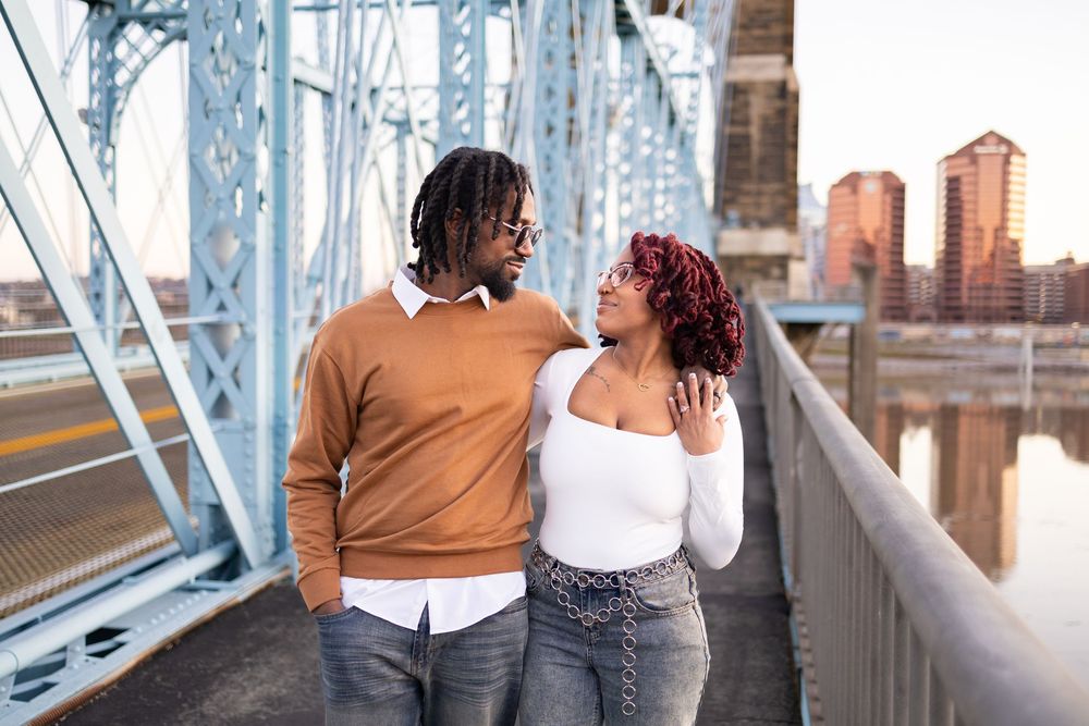 A couple smiles at each other while standing on Roebling bridge with city buildings in the background in Cincinnati Ohio.