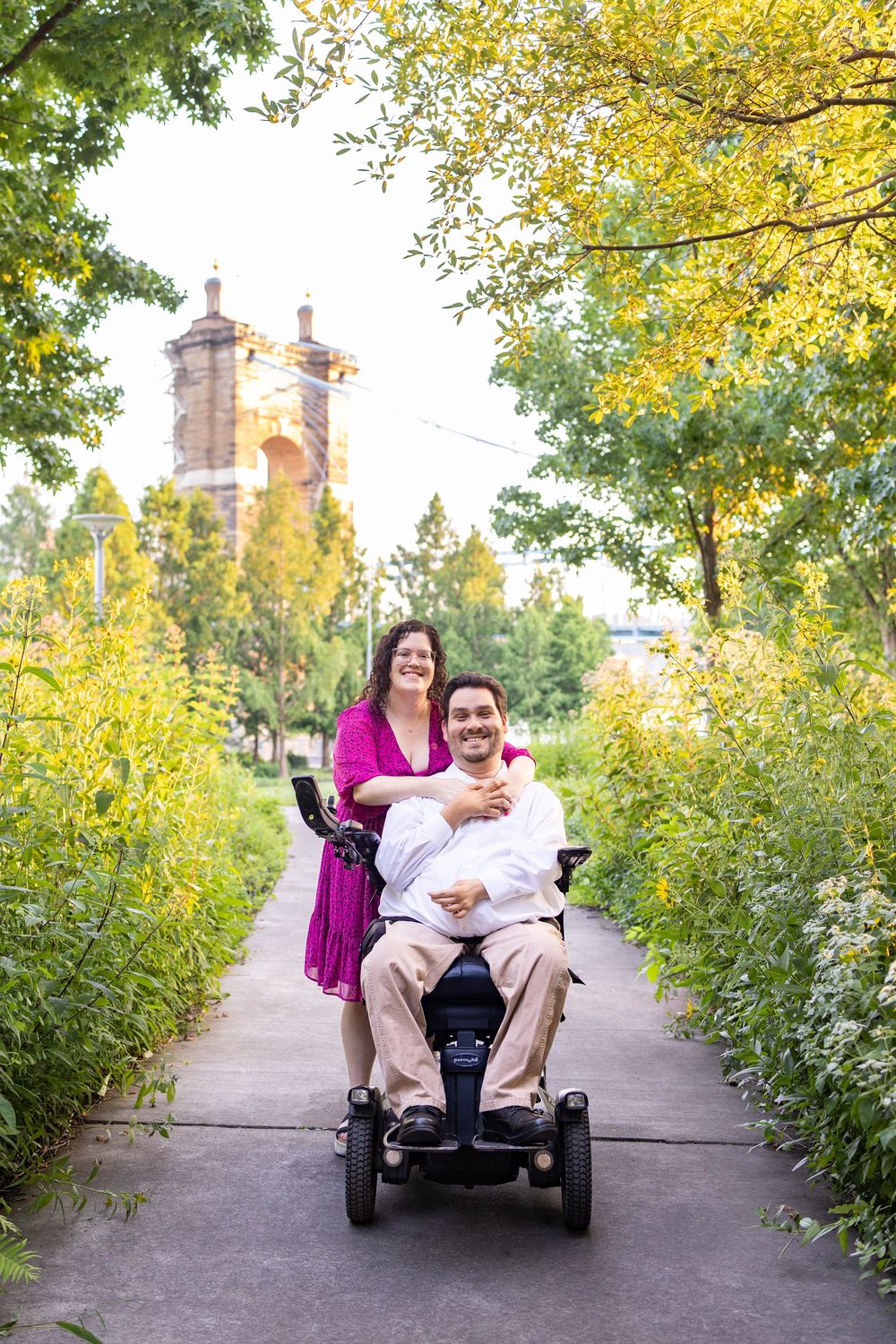 Smiling couple in park, man in power wheelchair with woman in pink dress behind him in front of Roebling Bridge in Smale Park Cincinnati.