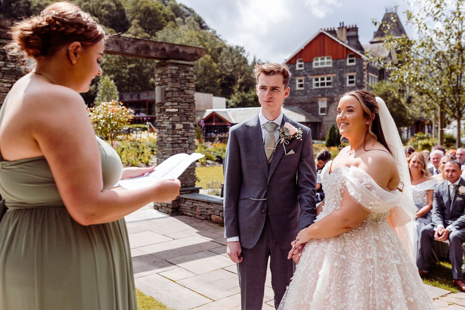 Bride and groom exchange vows outdoors at a scenic wedding ceremony, officiated by a woman in a sage green dress.