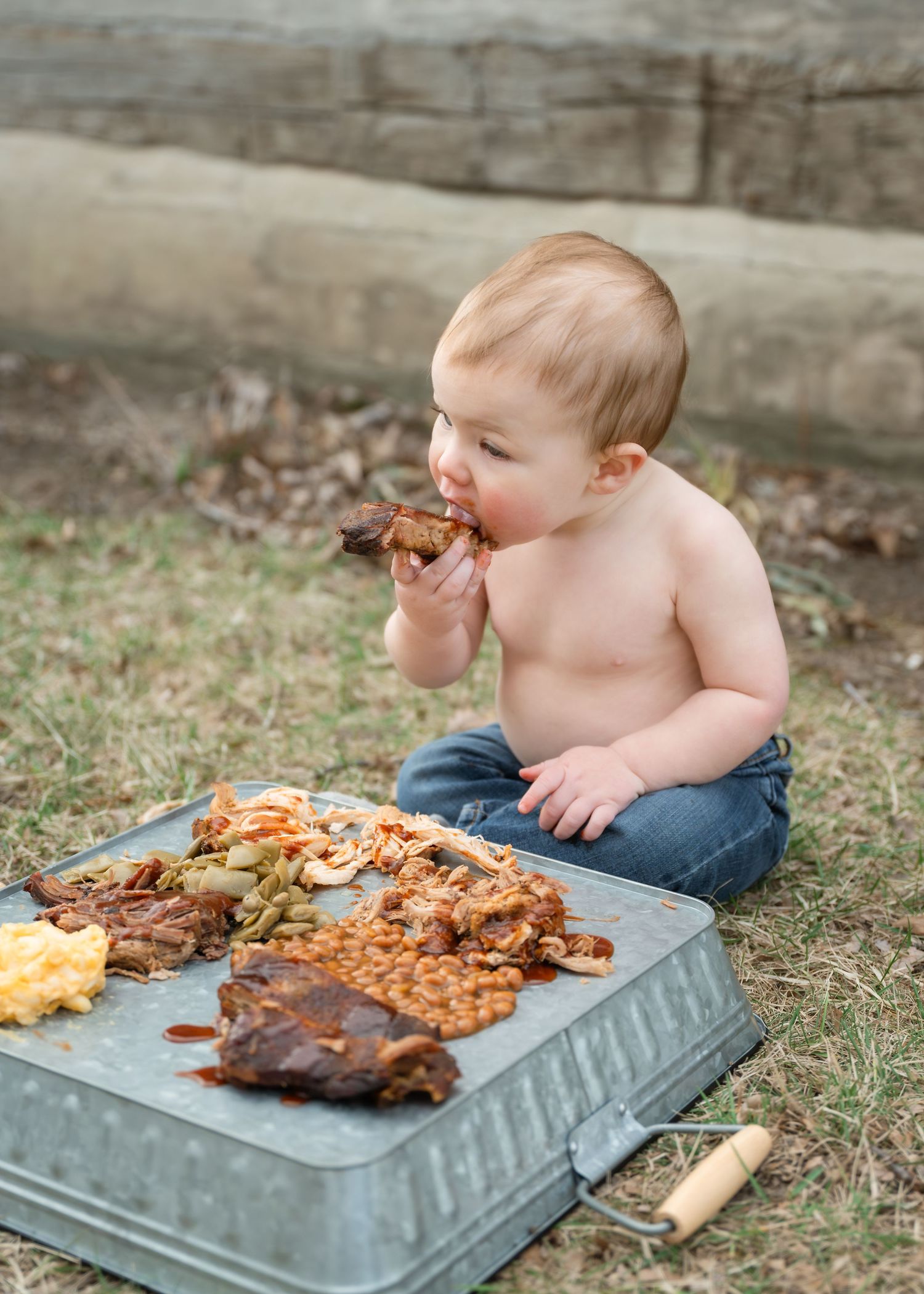 Shirtless baby sitting outdoors eating BBQ ribs from a metal tray filled with baked beans and sides.
