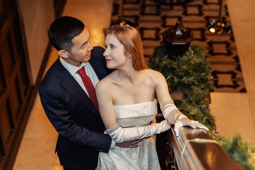 Elegant couple in formal wedding attire embracing on a balcony, groom in navy suit, bride in white strapless gown.