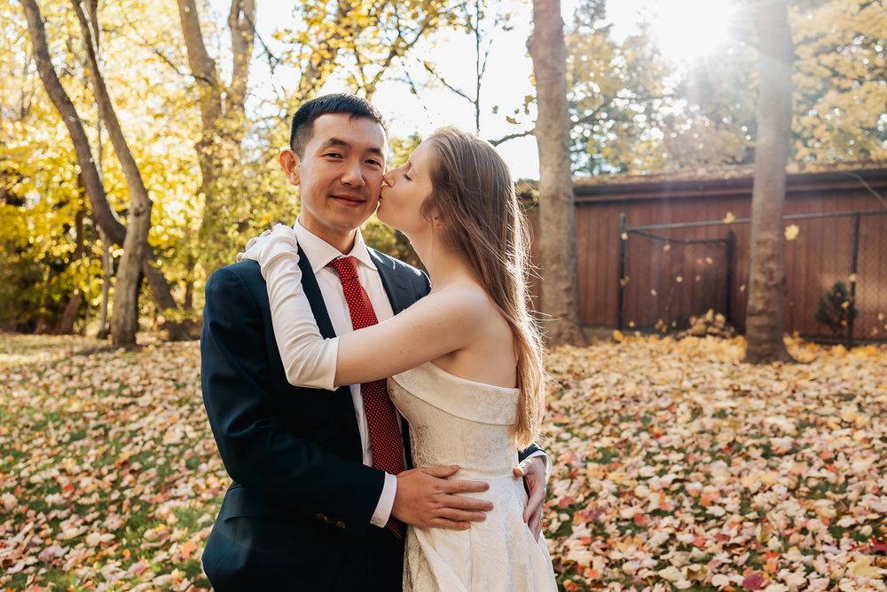 Couple in wedding attire embracing outdoors among golden autumn leaves, bride kissing groom on cheek in sunlit backyard.