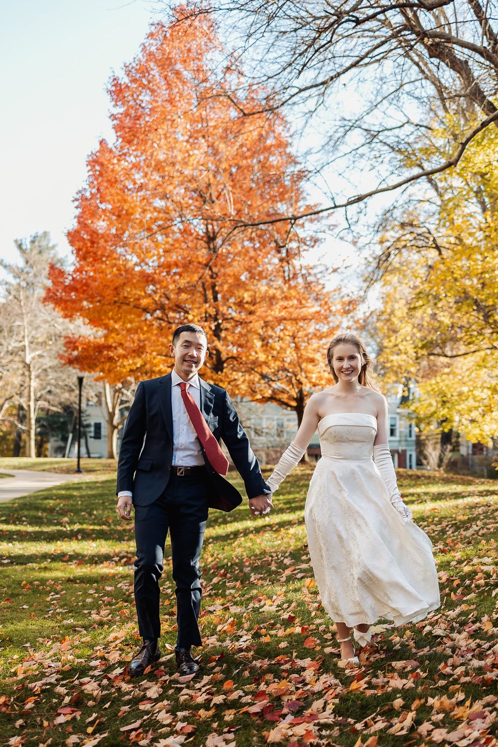 Joyful newlyweds holding hands and running through a park with vibrant autumn foliage on their wedding day.