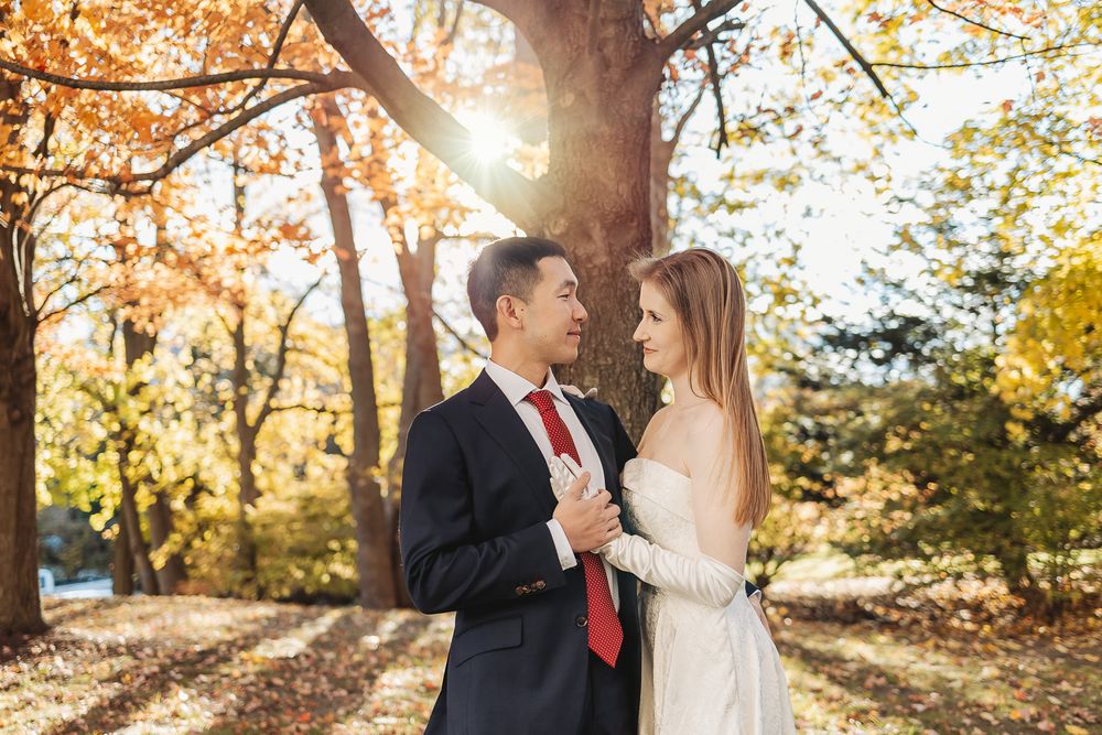 Couple in wedding attire embracing among autumn trees with golden sunlight in a romantic outdoor setting.