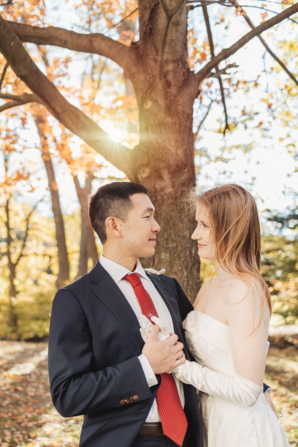 Couple in wedding attire embracing in autumn park, groom in navy suit with red tie, bride in white dress, sunlight through trees.