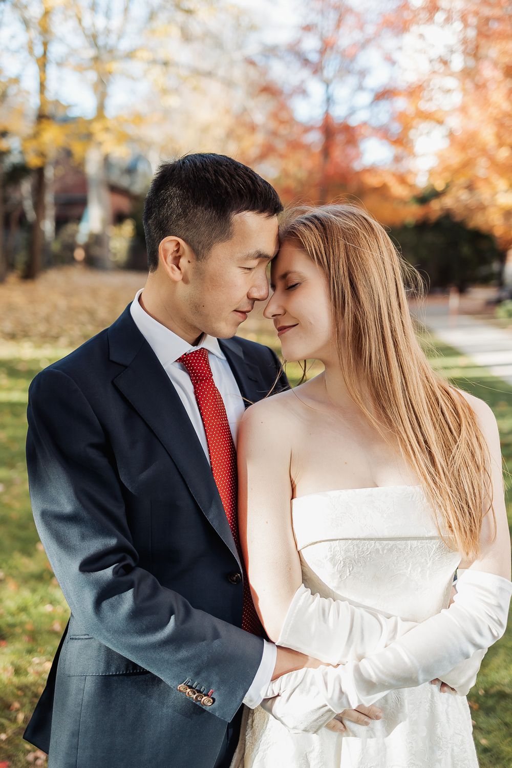 Couple in wedding attire embracing outdoors in autumn, groom in navy suit with red tie, bride in white dress.