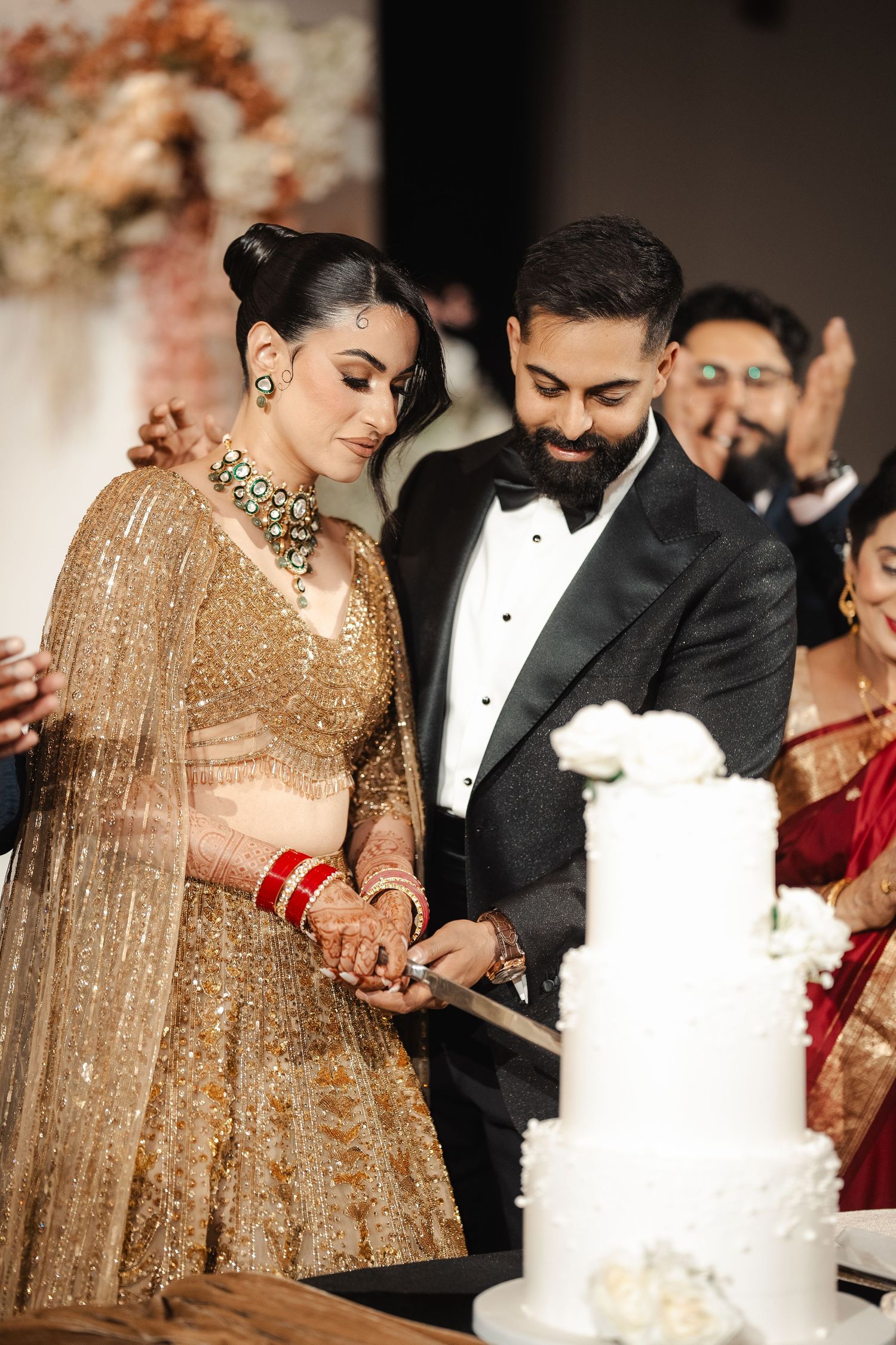 Indian bride in gold lehenga and groom in tuxedo cutting white wedding cake at elegant reception ceremony.