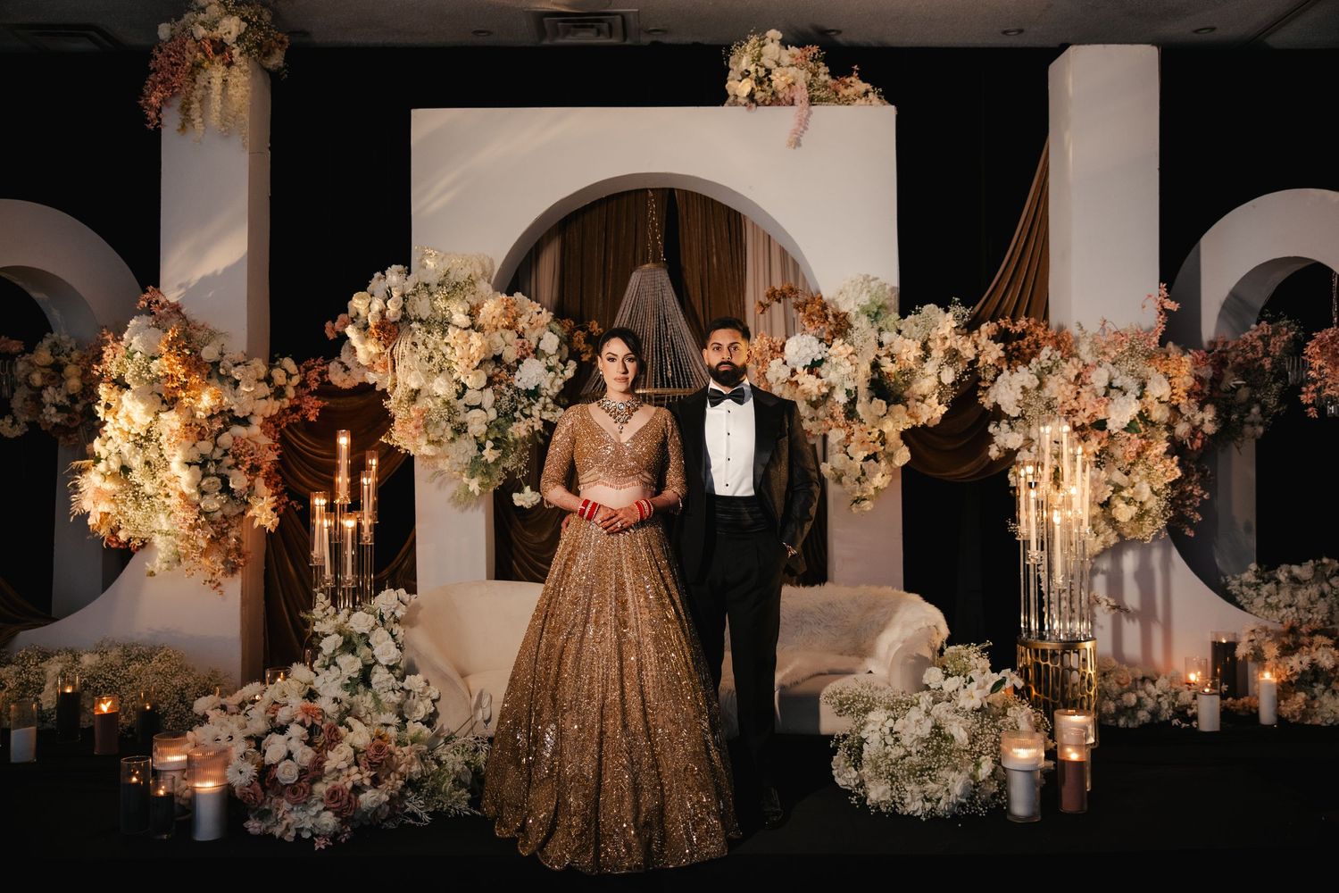 Elegant couple in gold lehenga and tuxedo posing before floral backdrop with candles at luxurious wedding reception.