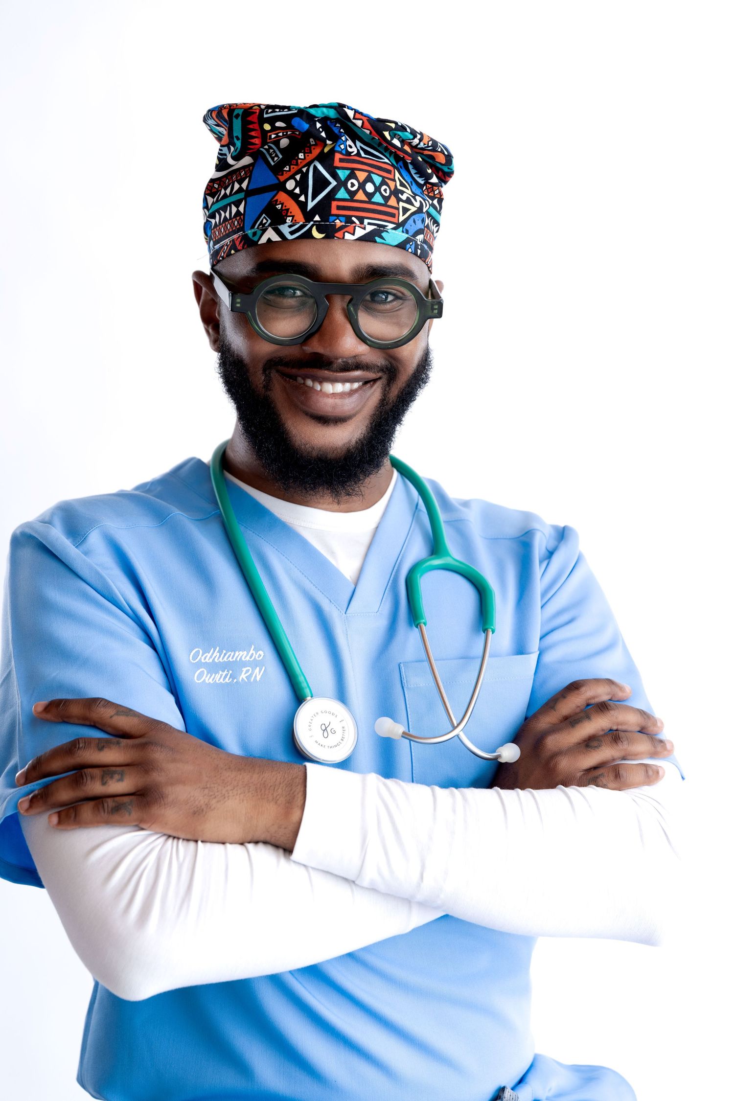 Smiling male nurse in blue scrubs, stethoscope, colorful African print cap, and glasses, standing with arms crossed.