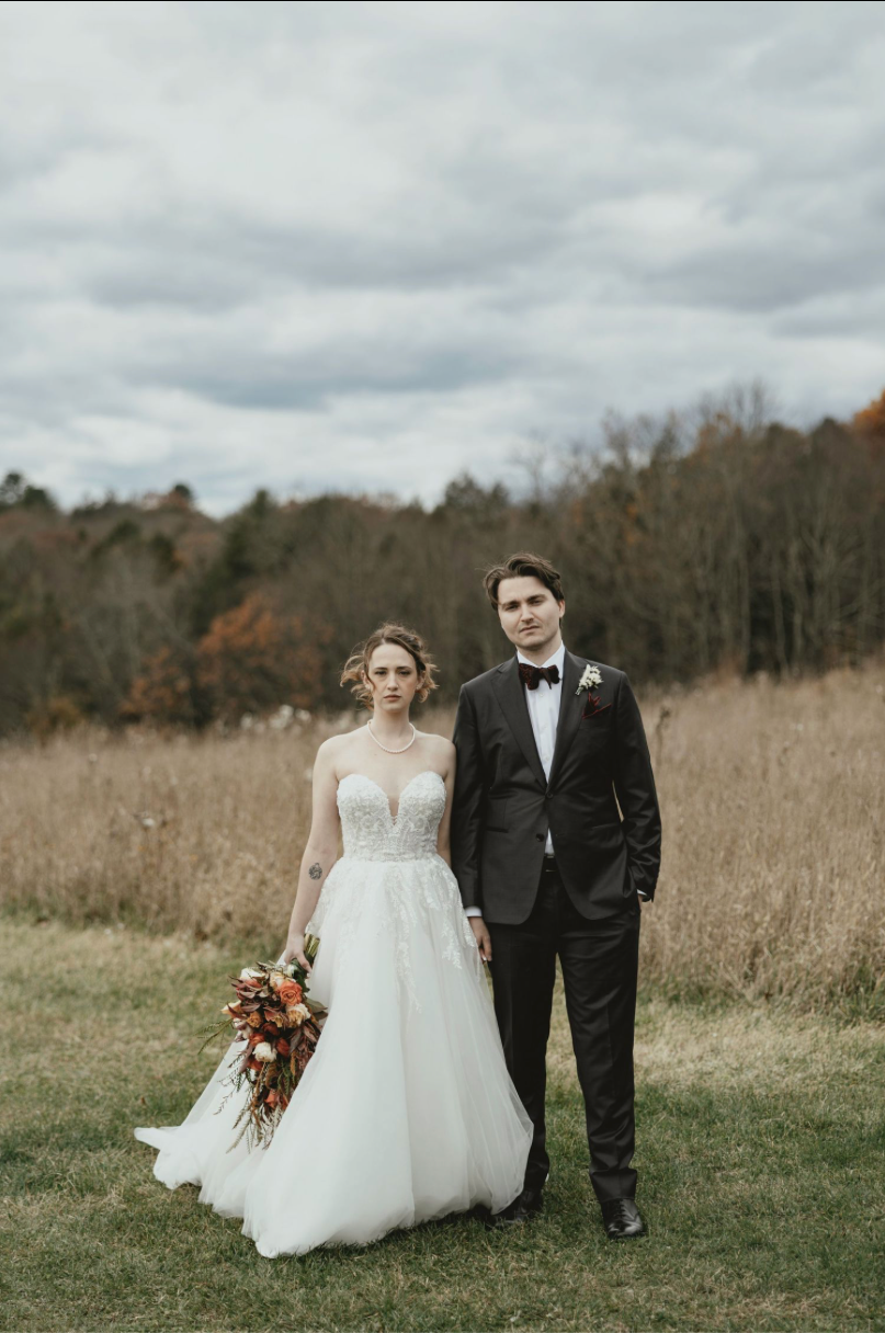 Bride in white lace gown holding bouquet stands in church aisle, bathed in natural light from stained glass window.