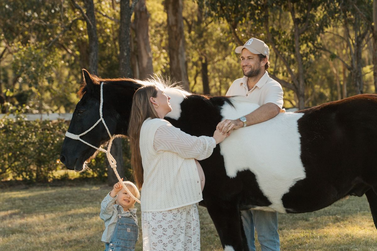 Couple bonding with black & white horse outdoors in a sunny, tree-lined paddock setting during a maternity photoshoot with a horse in Sydney