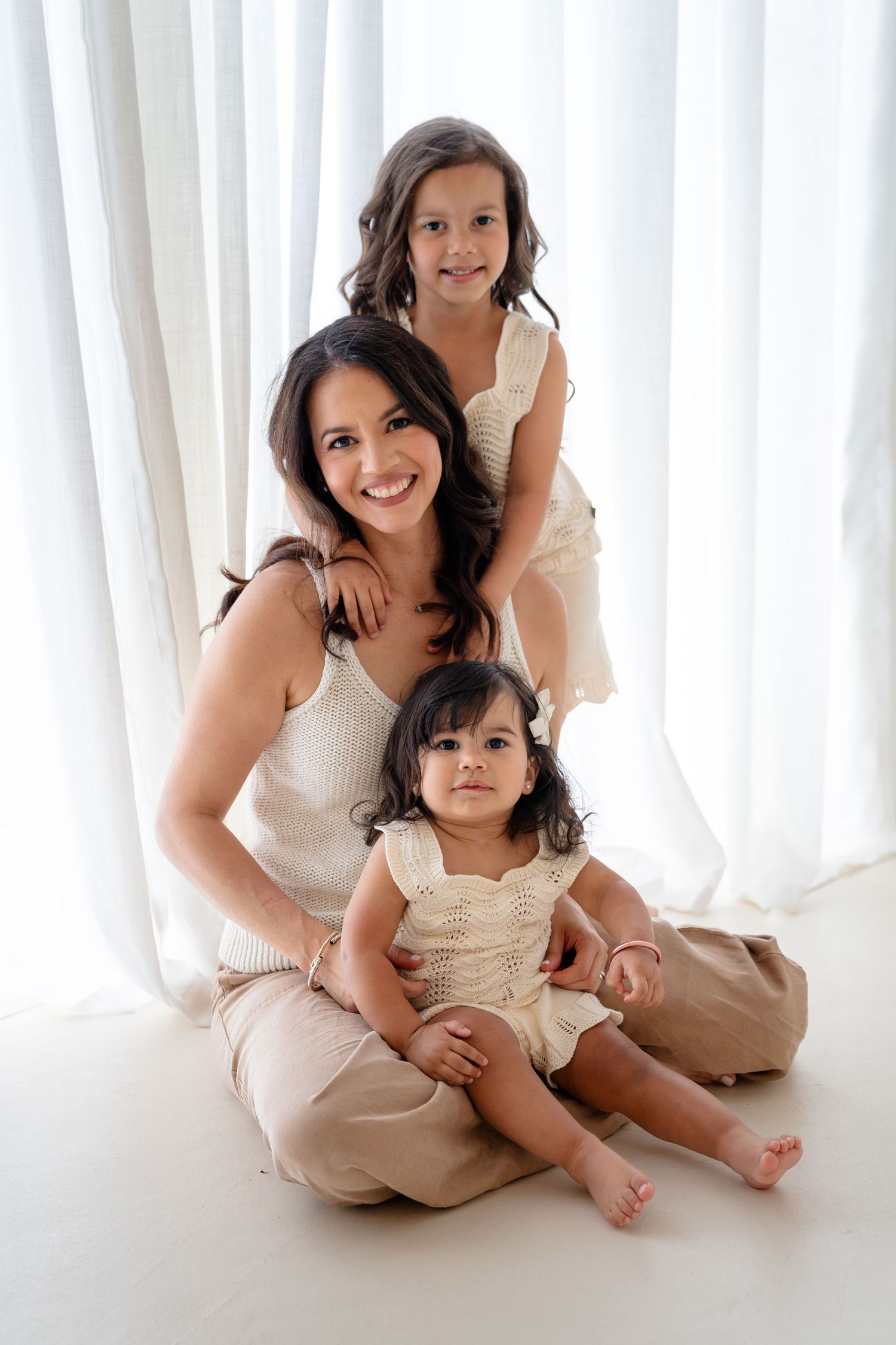 A smiling mother sits on the floor with two young daughters in matching cream outfits against white curtains.