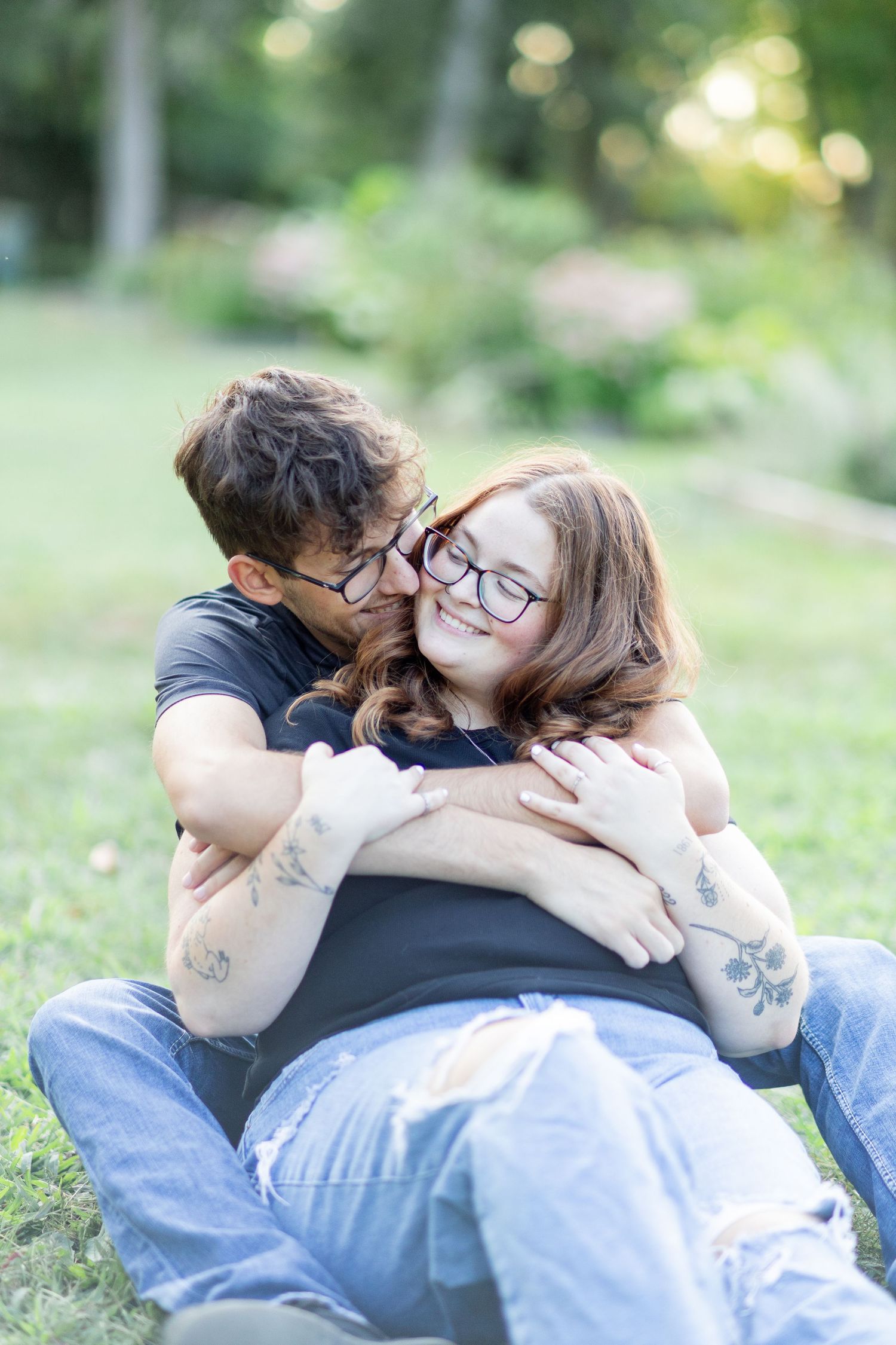 Couple sitting on grass, man hugging woman from behind, both wearing glasses, smiling, with tattoos visible outdoors.