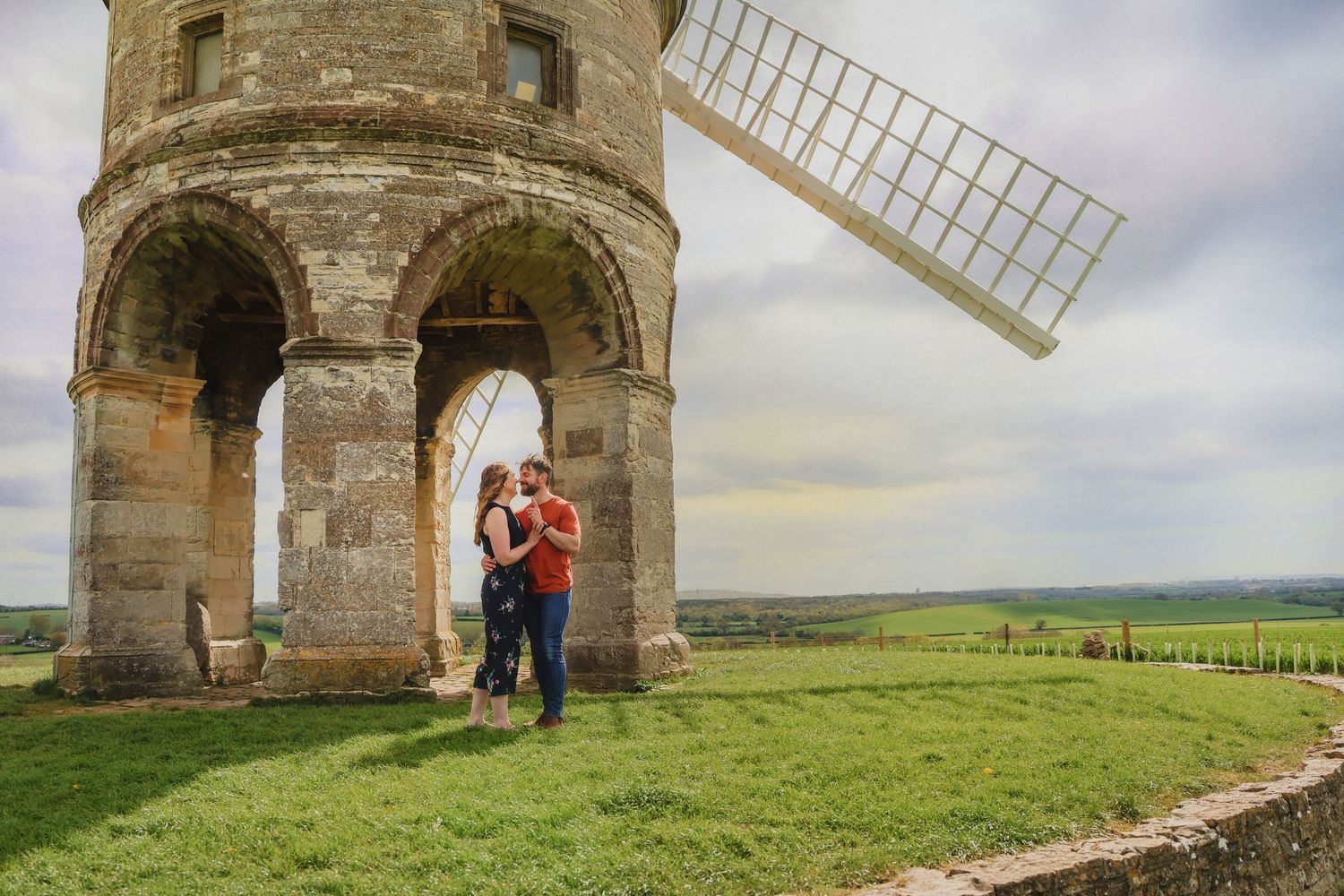 Couple embracing at the base of a historic stone windmill in a scenic countryside landscape.