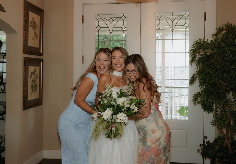 Bride in white gown poses with two bridesmaids holding bouquet near decorative glass front door.