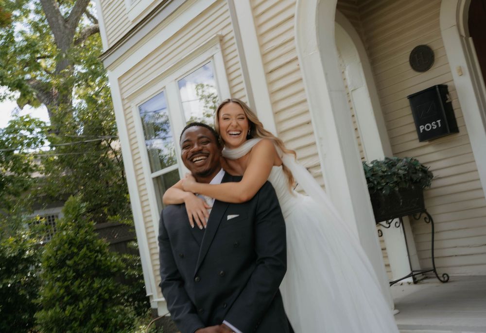 Joyful groom gives bride a piggyback ride while both laugh outside cream vintage building.