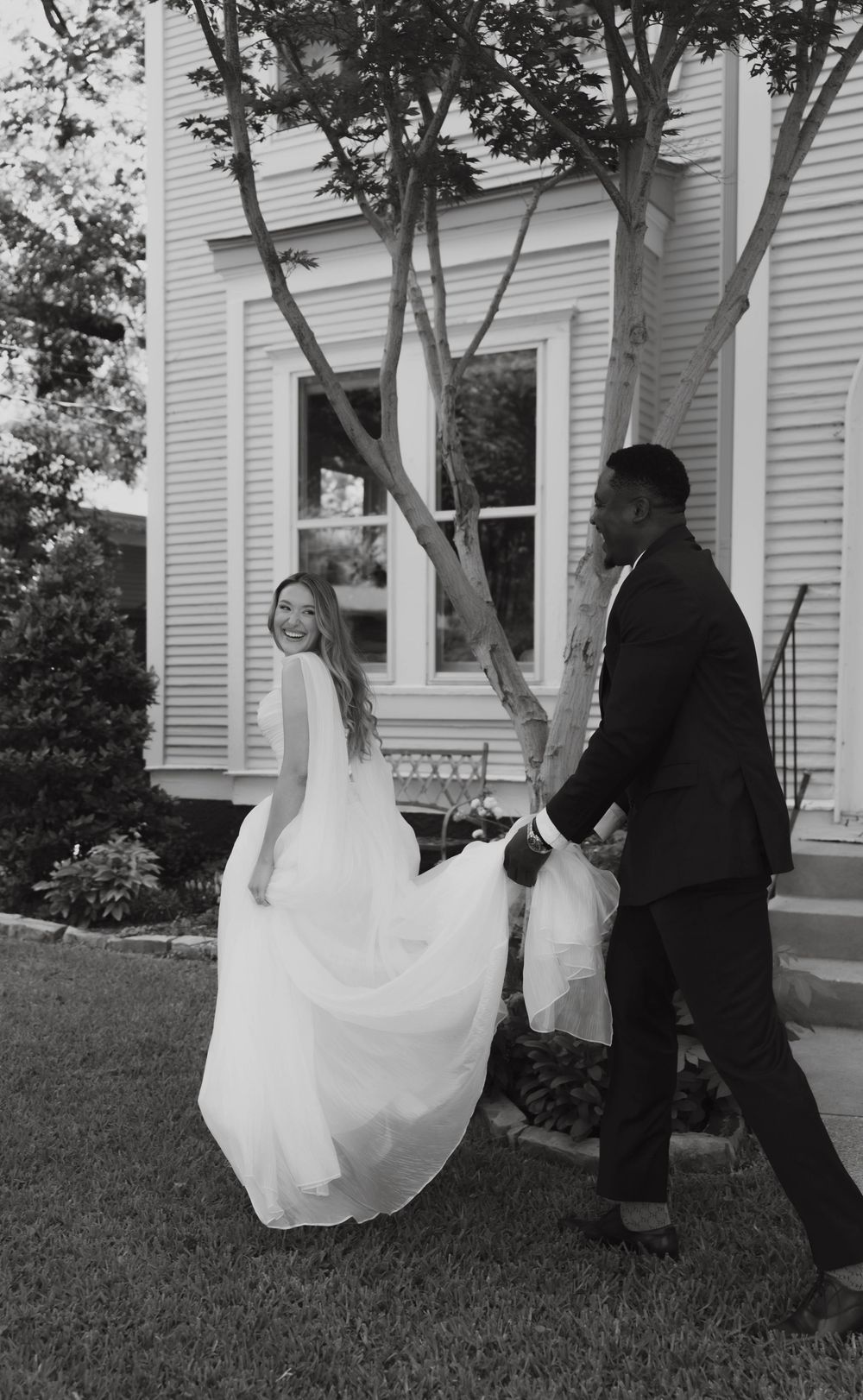 Black and white wedding photo of smiling bride and groom walking in garden beside a classic house.