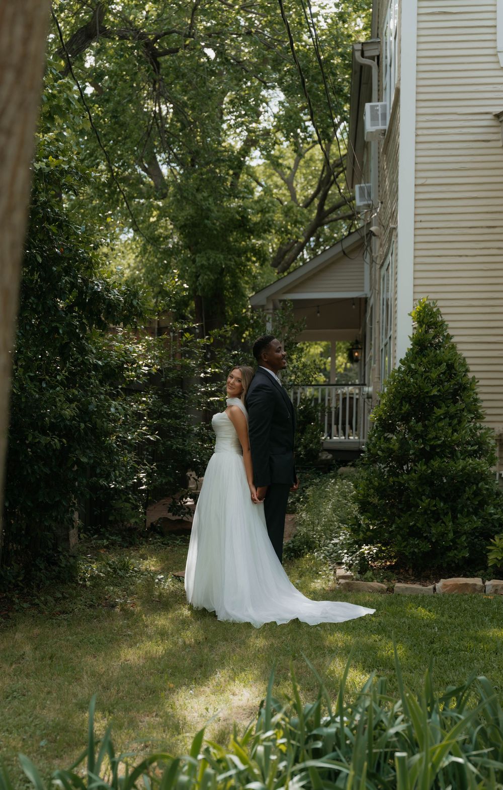 Bride and groom pose elegantly in a sunlit garden beside a historic home with tall trees.