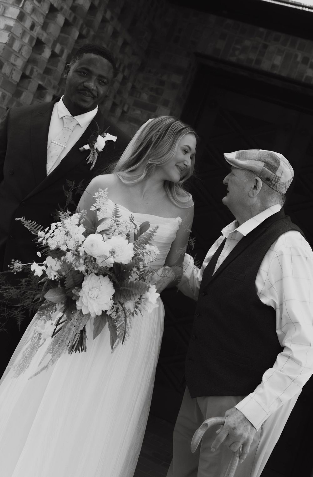 Black and white photo of bride with bouquet and groom greeting an elderly guest after the ceremony.