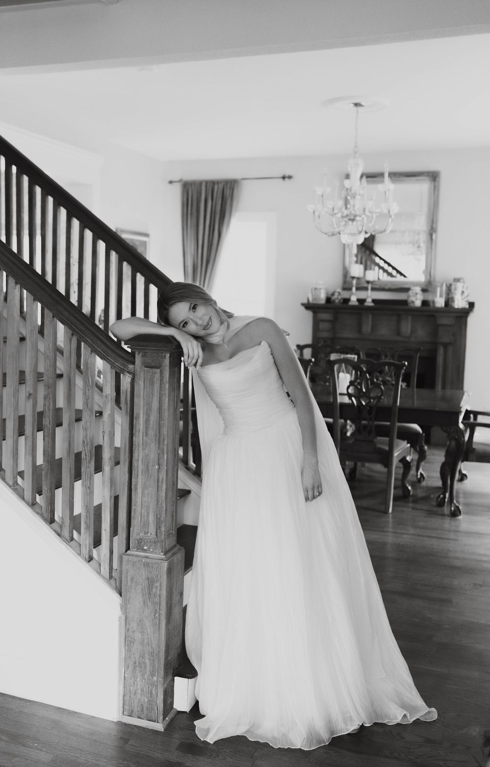 Elegant bride in strapless gown leans on staircase railing smiling in black and white photo.