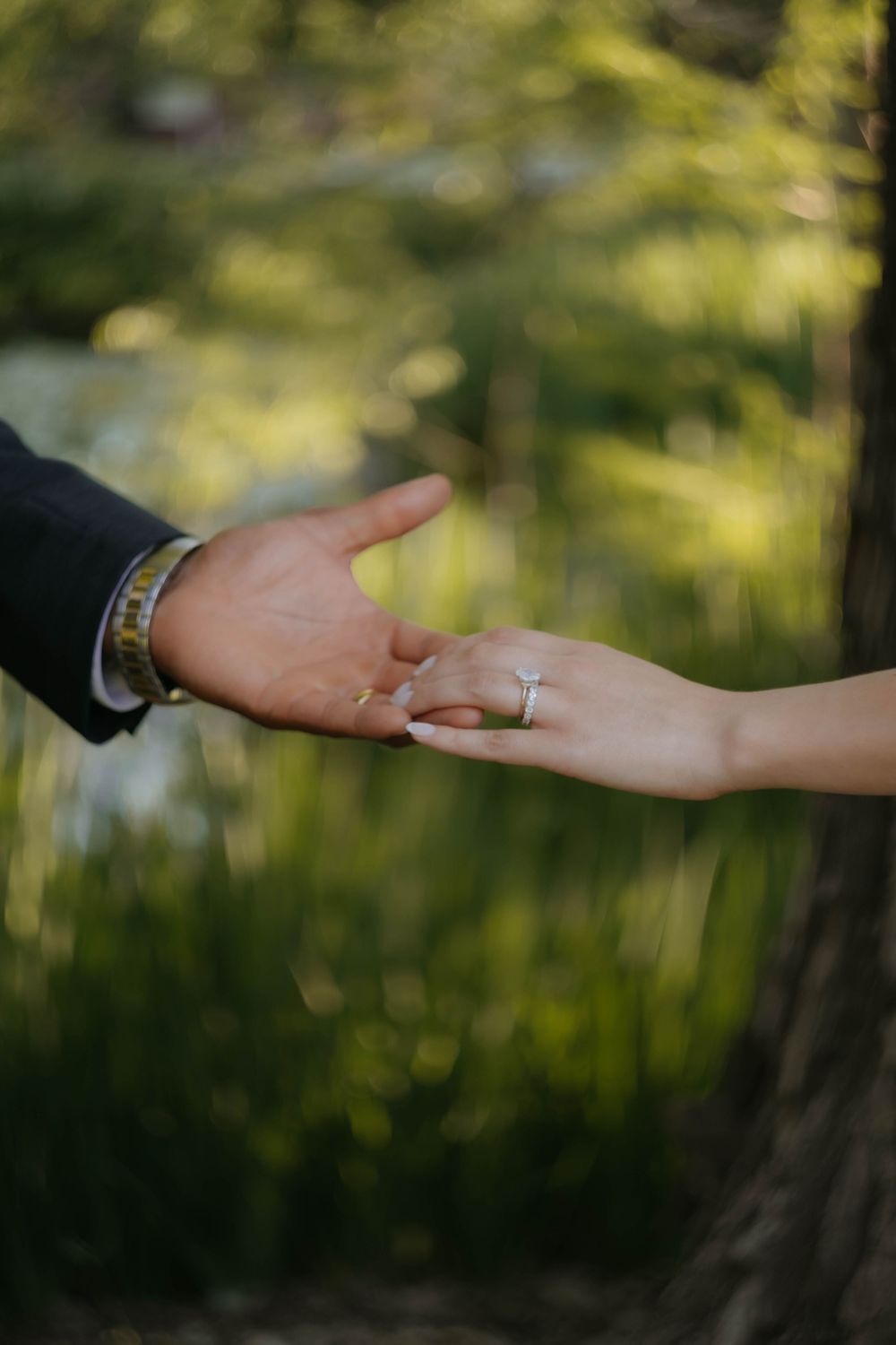 Close-up of bride and groom holding hands, showcasing wedding rings against a green bokeh background.