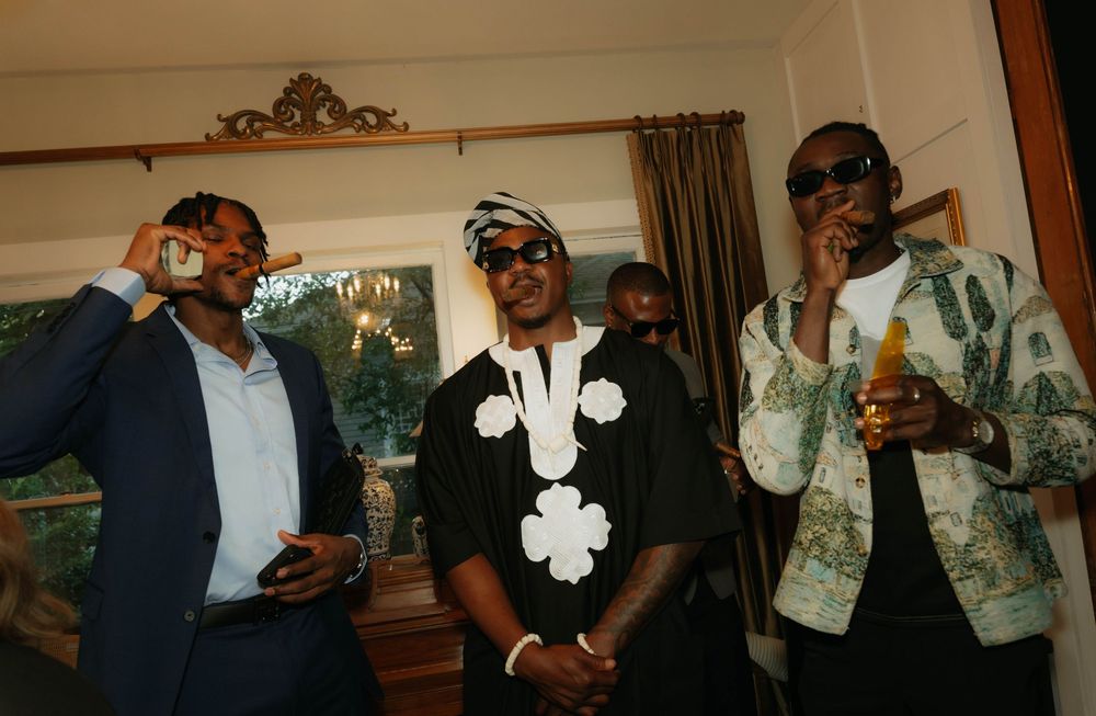 Three men in stylish attire celebrating indoors, one in a black floral outfit holding a drink.